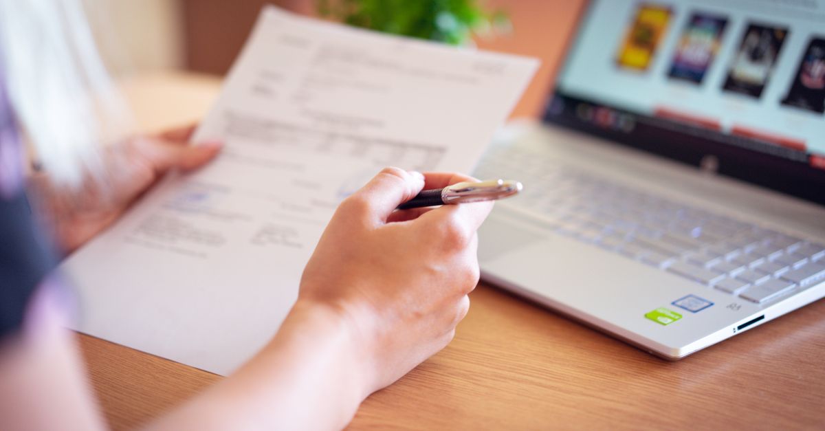 Close-up of a hand holding a pen and paper near a laptop, suggesting online shopping or document review. — illustrating create a reciept