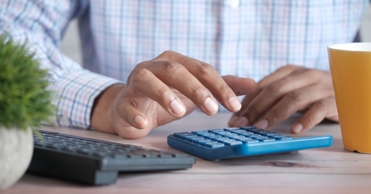 A person using a blue calculator on a wooden desk with a keyboard and plant nearby. — illustrating express expenses