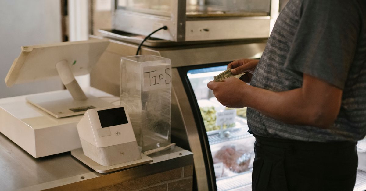 Unrecognizable male in casual clothes standing with money near cash register in grocery store while making purchase near glass showcase — illustrating make rece