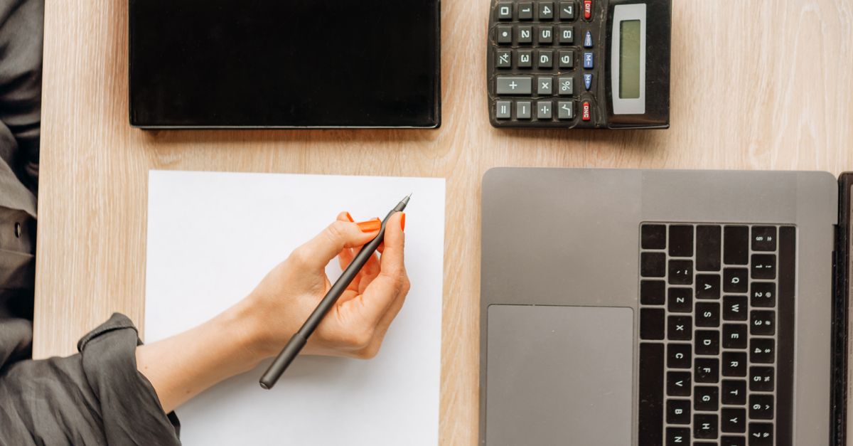 A person writing on paper surrounded by a laptop, tablet, and calculator on an office desk. — illustrating expenses receipt